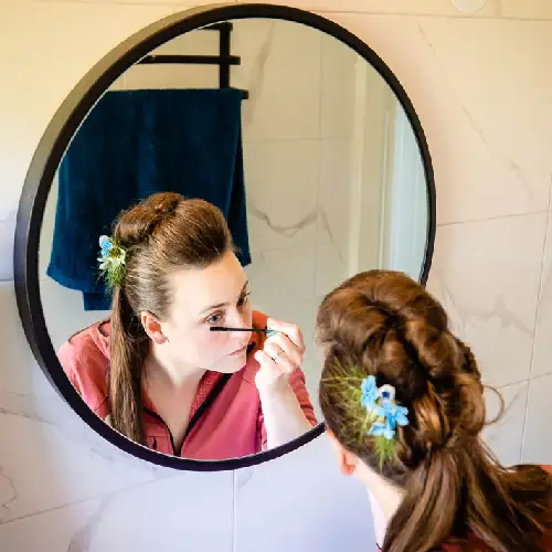 Een bruid in een roze ochtendjas kijkt geconcentreerd in een ronde badkamerspiegel terwijl ze haar mascara aanbrengt. Ze heeft een ingevlochten kapsel versierd met blauwe bloemetjes.