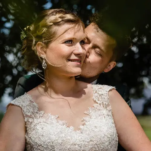 Een close-up van een bruid met een prachtig ingevlochten kapsel en een kanten trouwjurk, terwijl de bruidegom haar teder een kus op haar wang geeft tegen een achtergrond van onscherpe groene bomen.